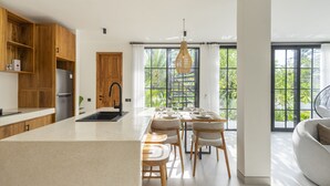 A modern kitchen with wooden cabinets, a central island, and a dining area with chairs and a table. - Vista Villa Ubud (Ubud)