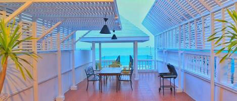 Área de refeições coberta ao ar livre, com mesa e cadeiras, com vista para o mar.