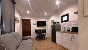 A compact living space with a sofa, a dining table with chairs, a kitchen area with cabinets and appliances, and a TV mounted on the wall.