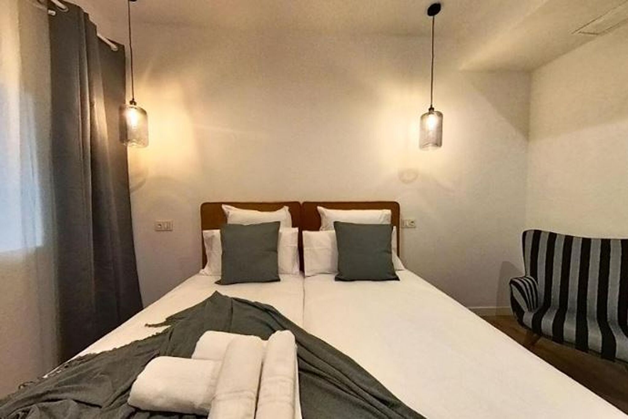 A double bed with white and green bedding, two hanging light fixtures, and a striped armchair.