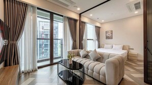 Een moderne hotelkamer met een bank, een bed, een glazen salontafel en grote ramen met gordijnen.