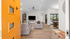A modern living room with a large TV, sofas, a ceiling fan, and a dining area with a table and chairs.