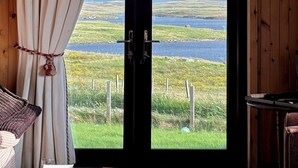 A room with a wooden paneled wall, a large window with curtains, a view of a lake, and a chair. - Snishival - a magical Hebridean retreat. Fantastic views of S Uist. Pet-friendly (Snishival)