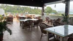 A covered outdoor seating area with wicker furniture and glass tables, overlooking a scenic view of palm trees and buildings.