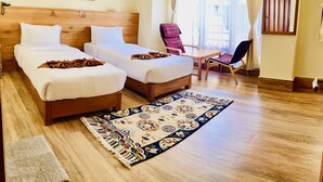 A hotel room with two single beds, a wooden headboard, a large window with curtains, a chair, a small table, and a colorful rug.