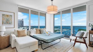 A modern living room with a large window, a sofa, a chair, a table, and a hanging light fixture. - Panoramic Vistas | Modern Apartment in Coconut (Miami)