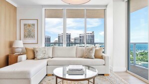 A modern living room with a sofa, a coffee table, and a view of a cityscape through large windows. - Panoramic Vistas | Modern Apartment in Coconut (Miami)