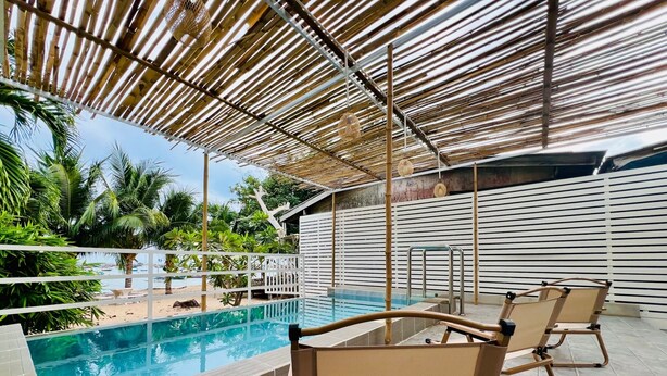 A pool area with a tiled deck, two lounge chairs, a pool with clear blue water, and a bamboo canopy overhead. - GAO Phala beachfront hostel & pool villa (Ban Chang)