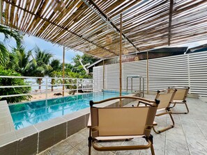Zona de piscina con terraza de baldosas, dos sillas de descanso, una piscina de agua cristalina y un toldo de bambú.