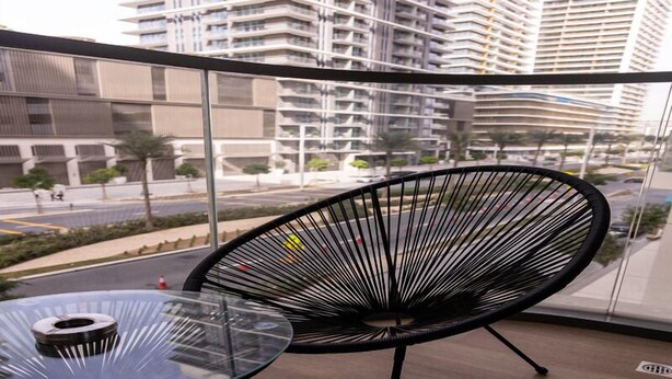 A modern balcony with a glass table and a black chair, offering a view of a cityscape with high-rise buildings and a road. - Tranquil 1BR Haven in the Heart of Vista (Dubai)