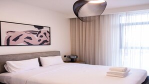 A modern hotel room with a large bed, a headboard, a wall art piece, a bedside table with a lamp, and a window with curtains. - Tranquil 1BR Haven in the Heart of Vista (Dubai)