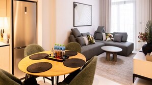 A modern living room with a grey sofa, a round dining table with chairs, a small coffee table, and a kitchen area with a refrigerator. - Tranquil 1BR Haven in the Heart of Vista (Dubai)