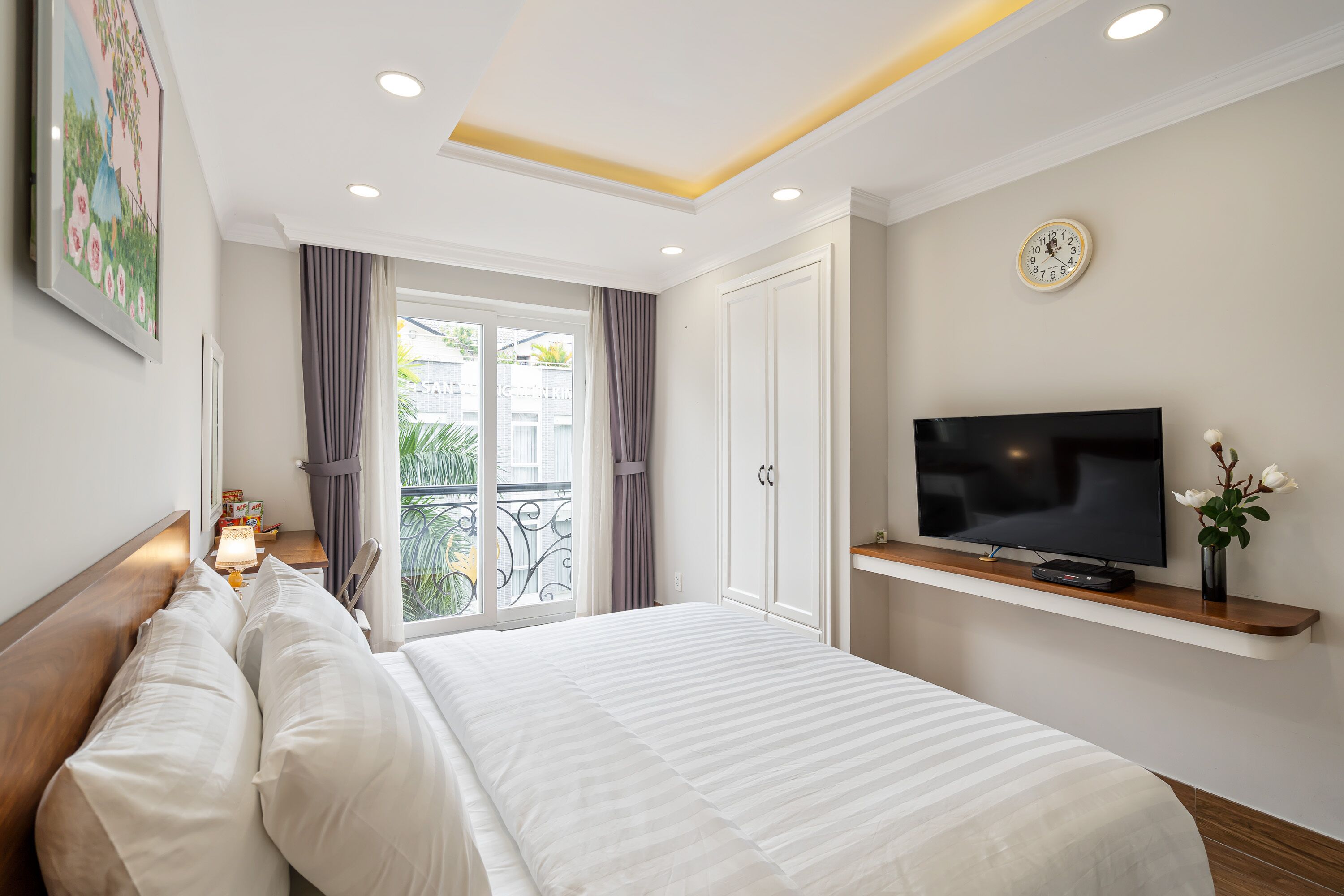 Quarto de hotel com uma cama grande, uma TV em uma estante, um relógio na parede e uma varanda com cortinas.