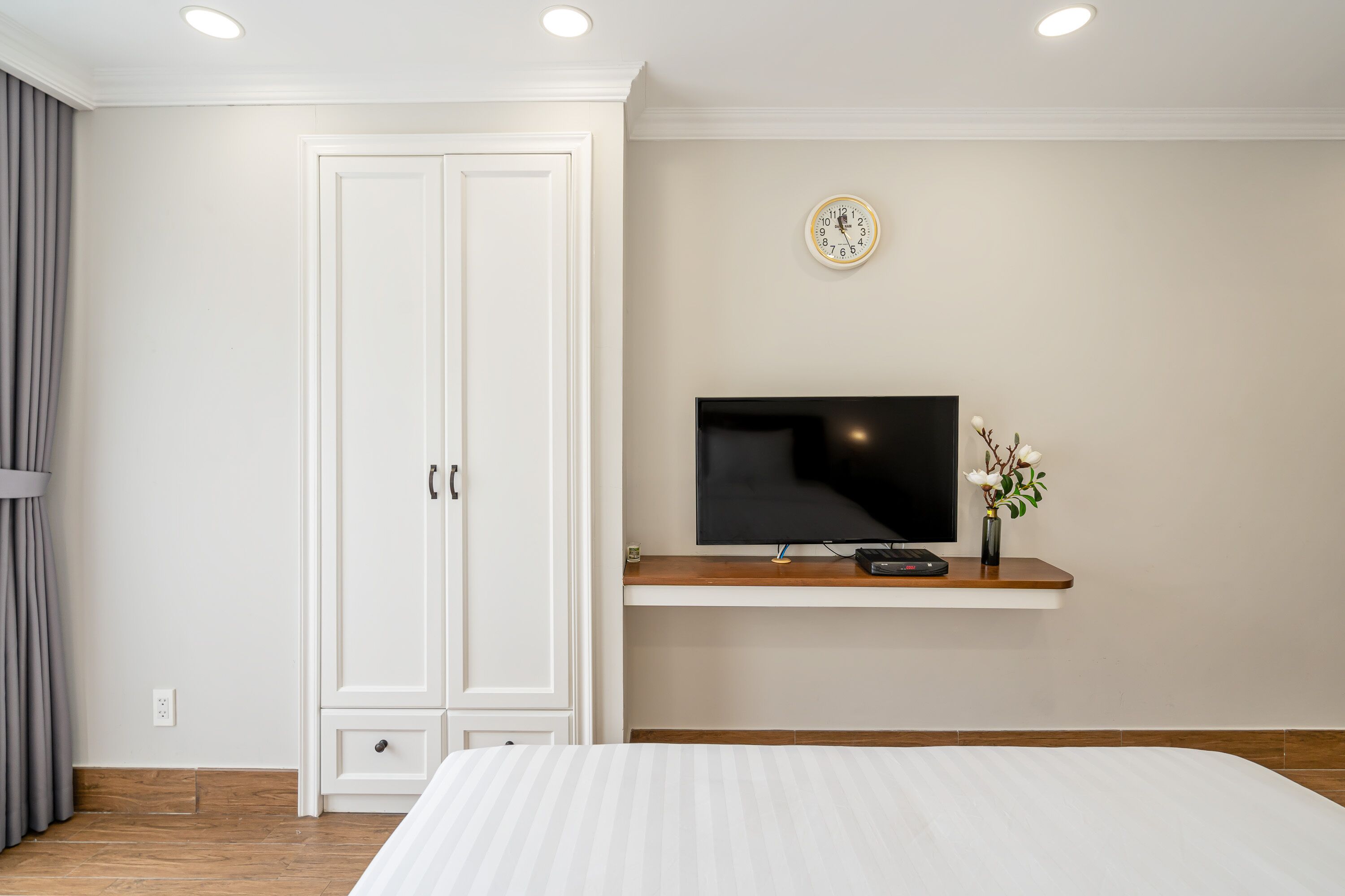 Een slaapkamer met een bed, een televisie op een plank, een kledingkast en een klok aan de muur.