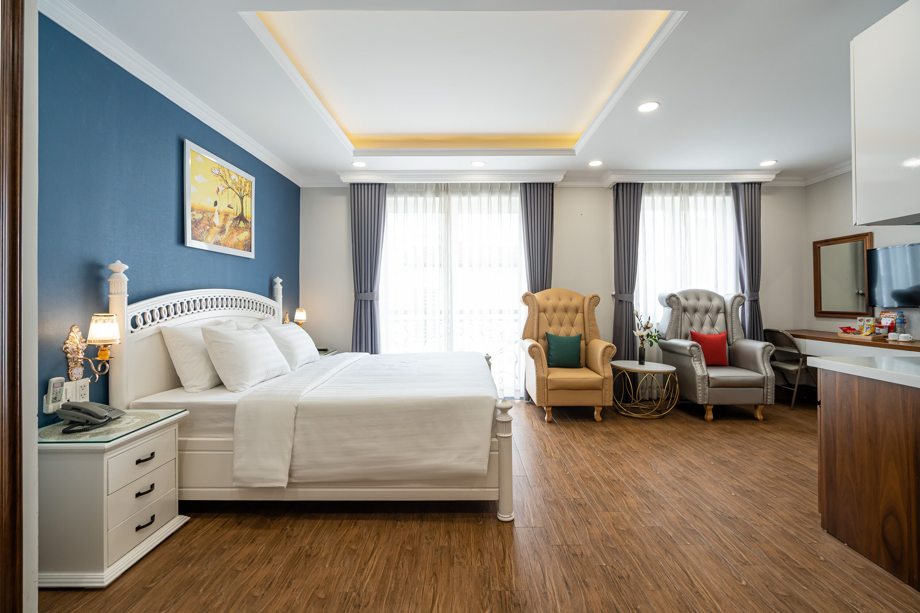 A hotel room with a large bed, a desk, two armchairs, a small side table, and a window with curtains.