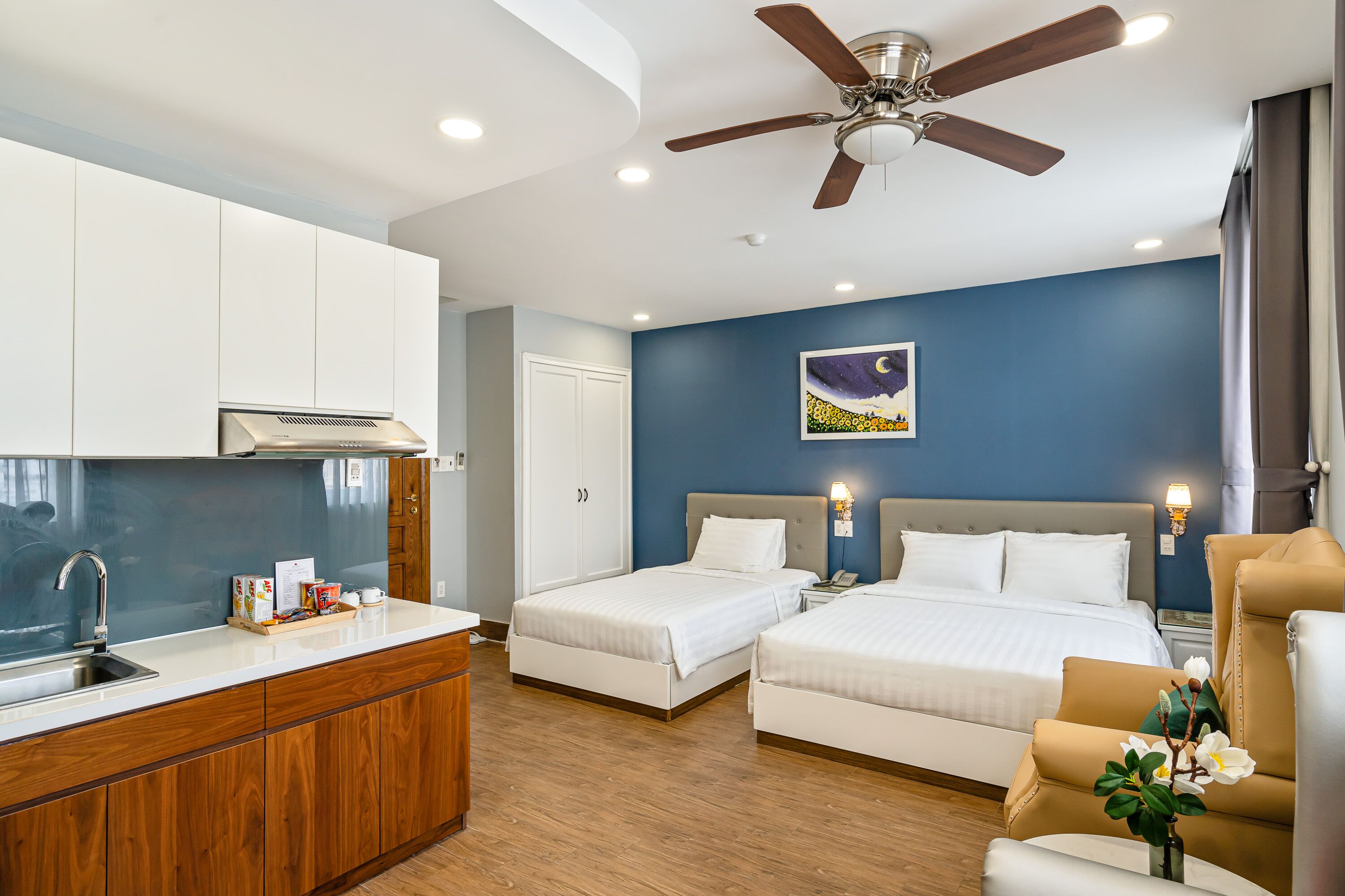 A hotel room with a kitchenette, two beds, a ceiling fan, and a nightstand with a lamp.