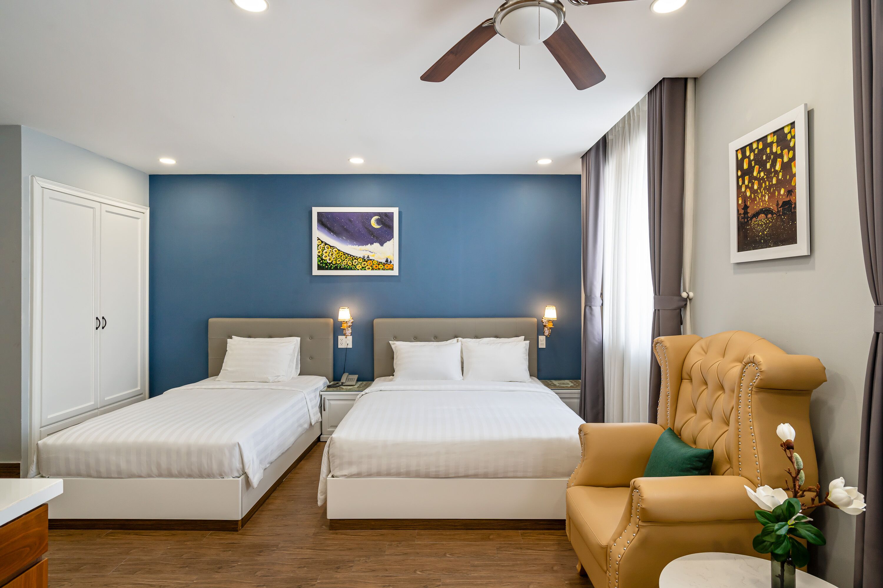 A hotel room with two beds, a ceiling fan, artwork on the walls, and a chair with a vase of flowers.