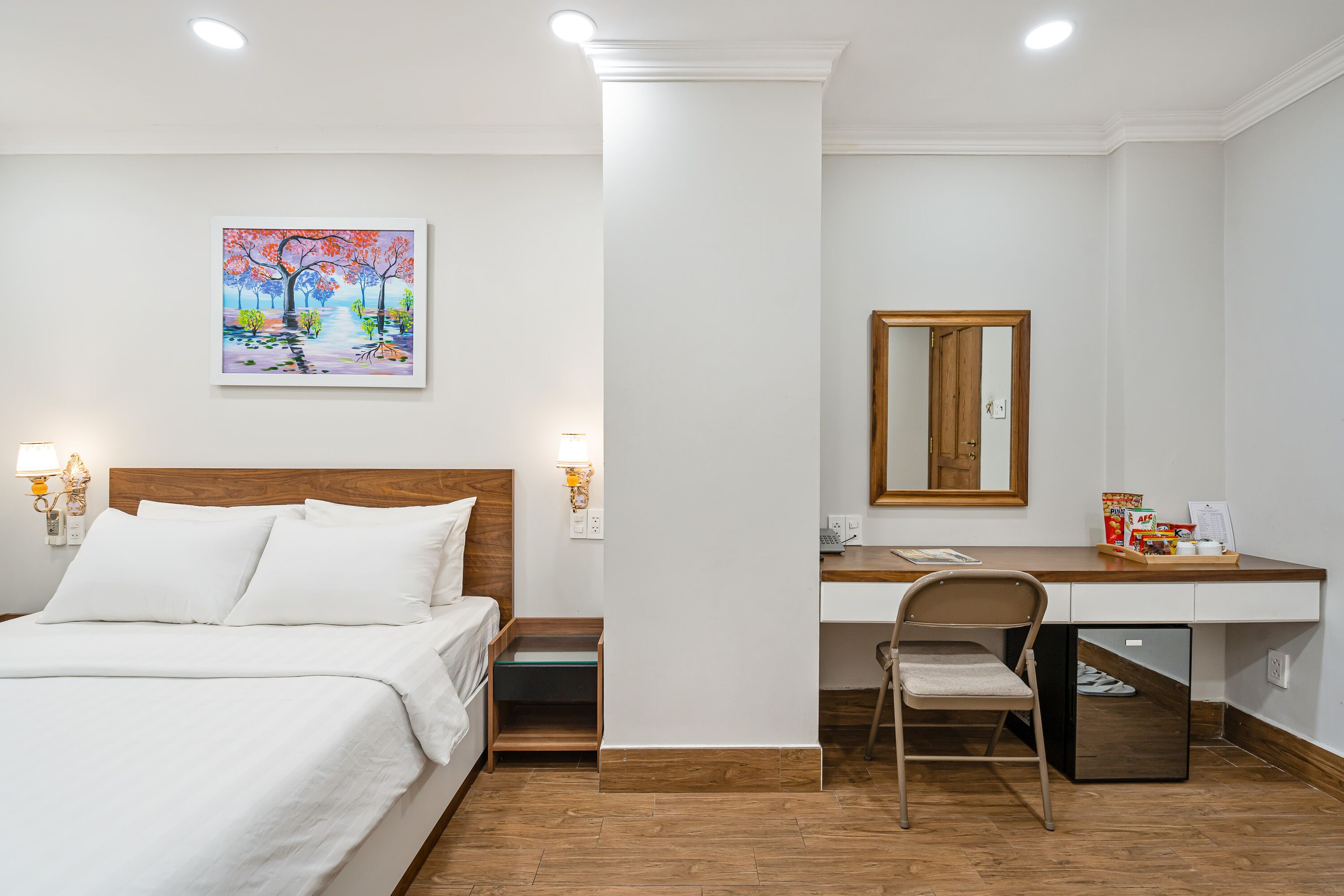 A hotel room with a bed, a desk, a chair, a small refrigerator, and a painting on the wall.