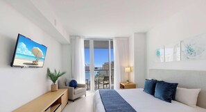 A hotel room with a bed, a TV, a desk, a chair, a balcony with a view of the city, and a window with curtains.