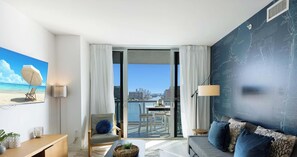 A modern hotel room with a balcony, a large window, a sofa, a coffee table, and a painting of a beach scene.