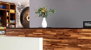A reception area with a wooden reception desk, a white vase with green foliage, and a wall with a circular design. - Hampton Inn & Suites By Hilton Pittsboro (Pittsboro)