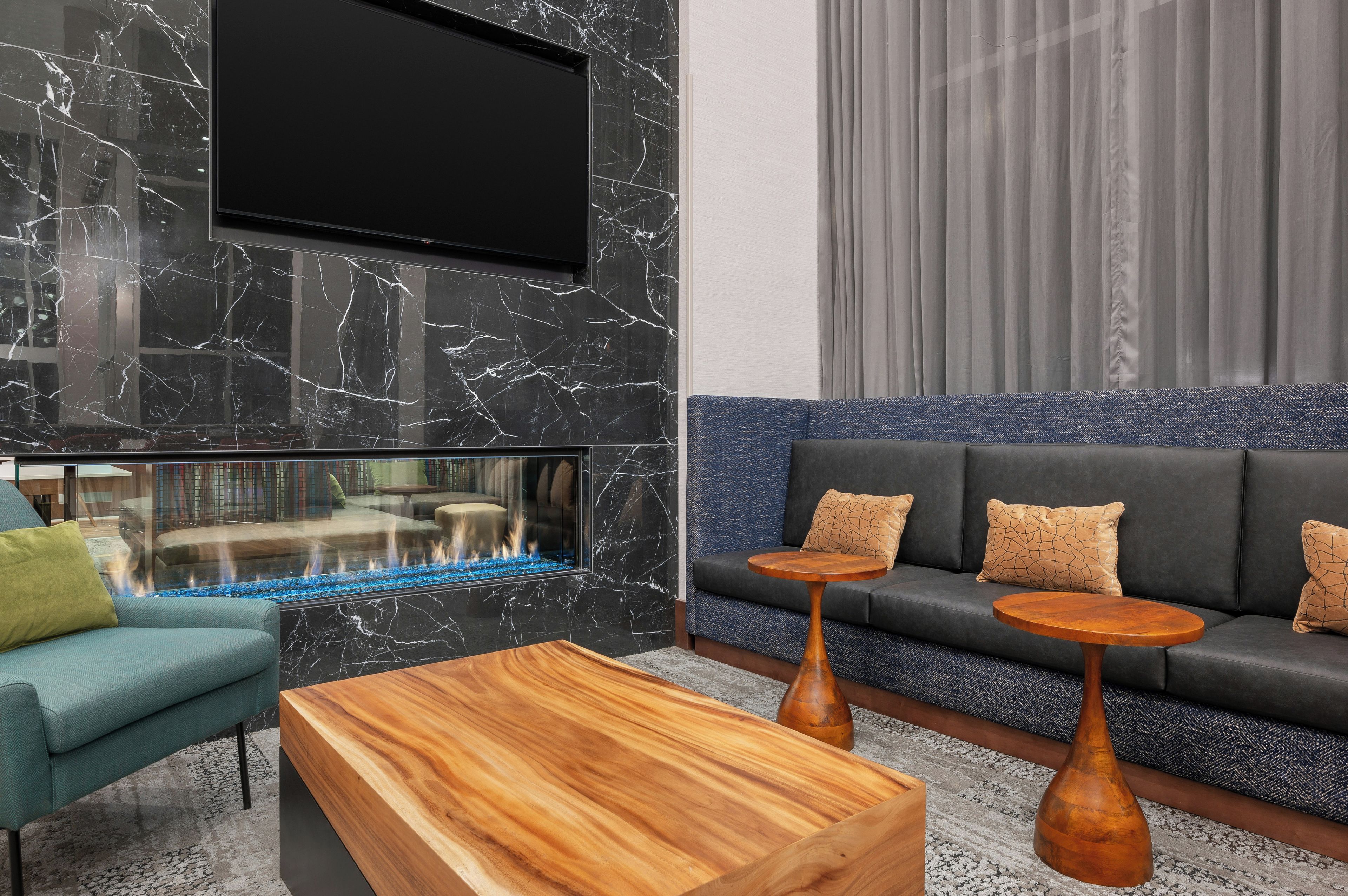 A modern living room with a black marble wall, a flat-screen TV, a glass fireplace, a grey sofa with patterned cushions, and wooden side tables.
