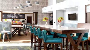 A modern hotel lobby with a long reception desk, teal chairs, and a large TV on the wall. - Hampton Inn & Suites By Hilton Pittsboro (Pittsboro)