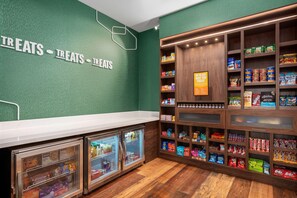 A snack bar with a green wall, wooden shelves stocked with various snacks, and two glass-fronted refrigerators. - Hampton Inn & Suites By Hilton Pittsboro (Pittsboro)