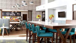 A modern hotel lobby with a long reception desk, teal chairs, and a large TV on the wall. - Hampton Inn & Suites By Hilton Pittsboro (Pittsboro)