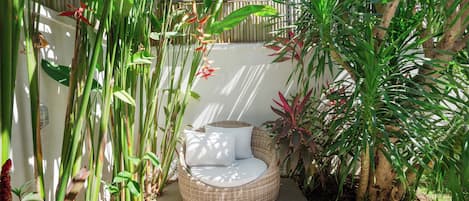 Un jardín con una silla de mimbre y una pequeña mesa, rodeado de diversas plantas tropicales.