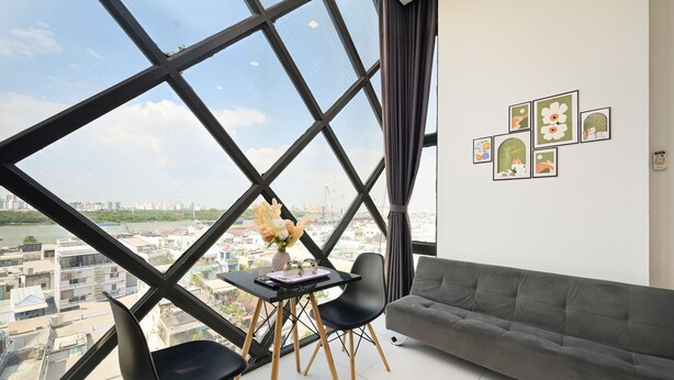 A modern room with a large window, a gray sofa, a small table with chairs, and wall art. - Huyen Apartment (Ho Chi Minh City)