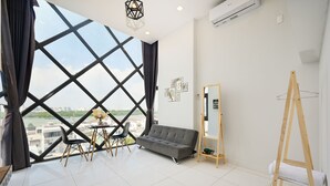 A modern room with a large window, a grey sofa, a wooden easel, and a small table with chairs. - Huyen Apartment (Ho Chi Minh City)