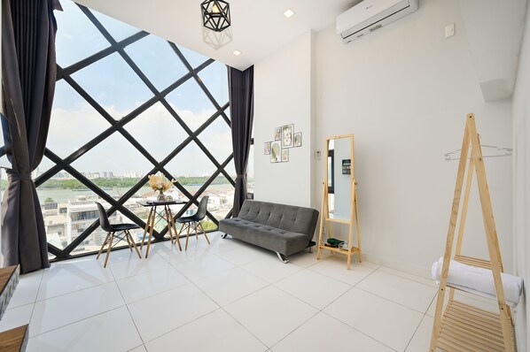 A modern room with a large window, a grey sofa, a wooden easel, and a small table with chairs. - Huyen Apartment (Ho Chi Minh City)