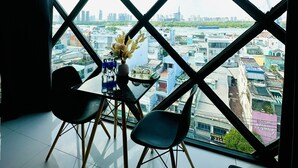 A room with a large window offering a city view, a small table with a vase of flowers, and two chairs. - Huyen Apartment (Ho Chi Minh City)