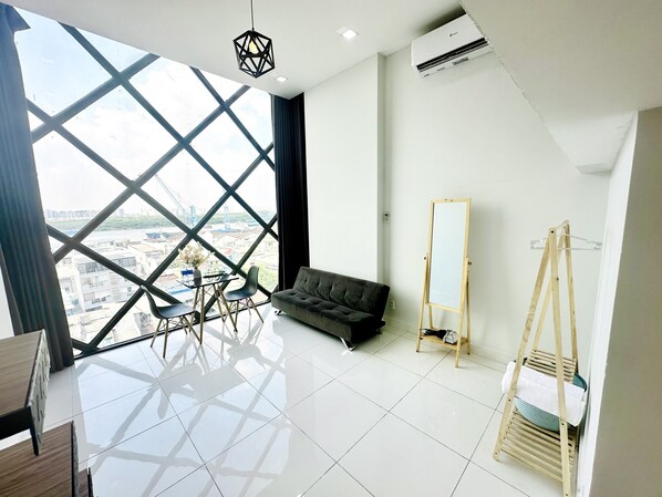A modern room with a large window, a sofa, a small table with chairs, a wooden easel, and a hanging rack. - Huyen Apartment (Ho Chi Minh City)