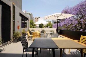 Un espace extérieur aménagé avec des tables et des chaises, entouré de plantes en pot et surmonté d’un grand parasol.