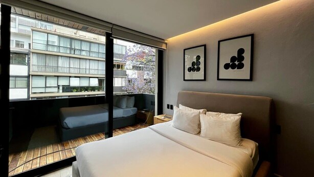 Ein modernes Hotelzimmer mit einem großen Bett, einer Sitzecke mit Sofa und Tisch sowie Blick auf ein Gebäude durch eine Glastür.