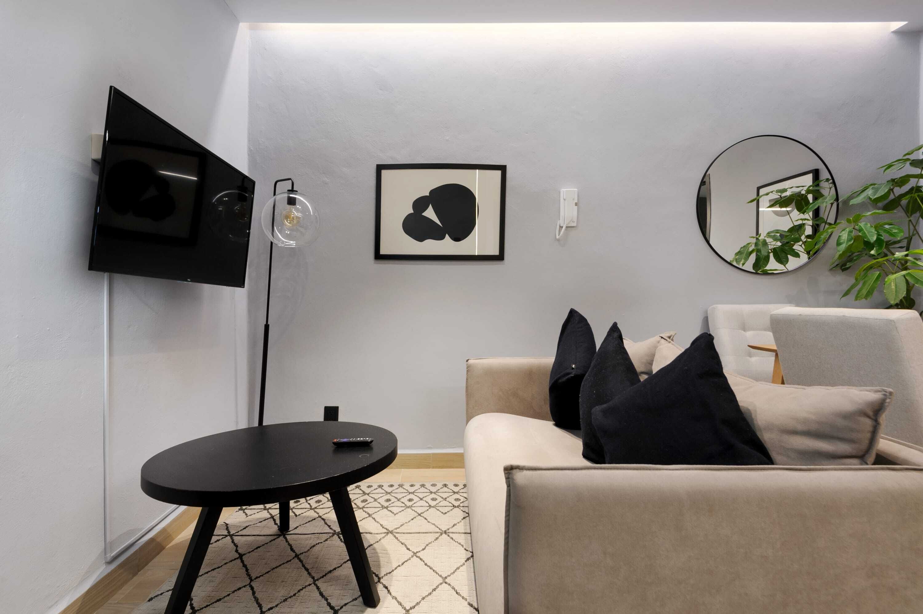 Een moderne woonkamer met een bank, een klein rond tafeltje, een vloerlamp, een flatscreen televisie en lijstwerken aan de muur.