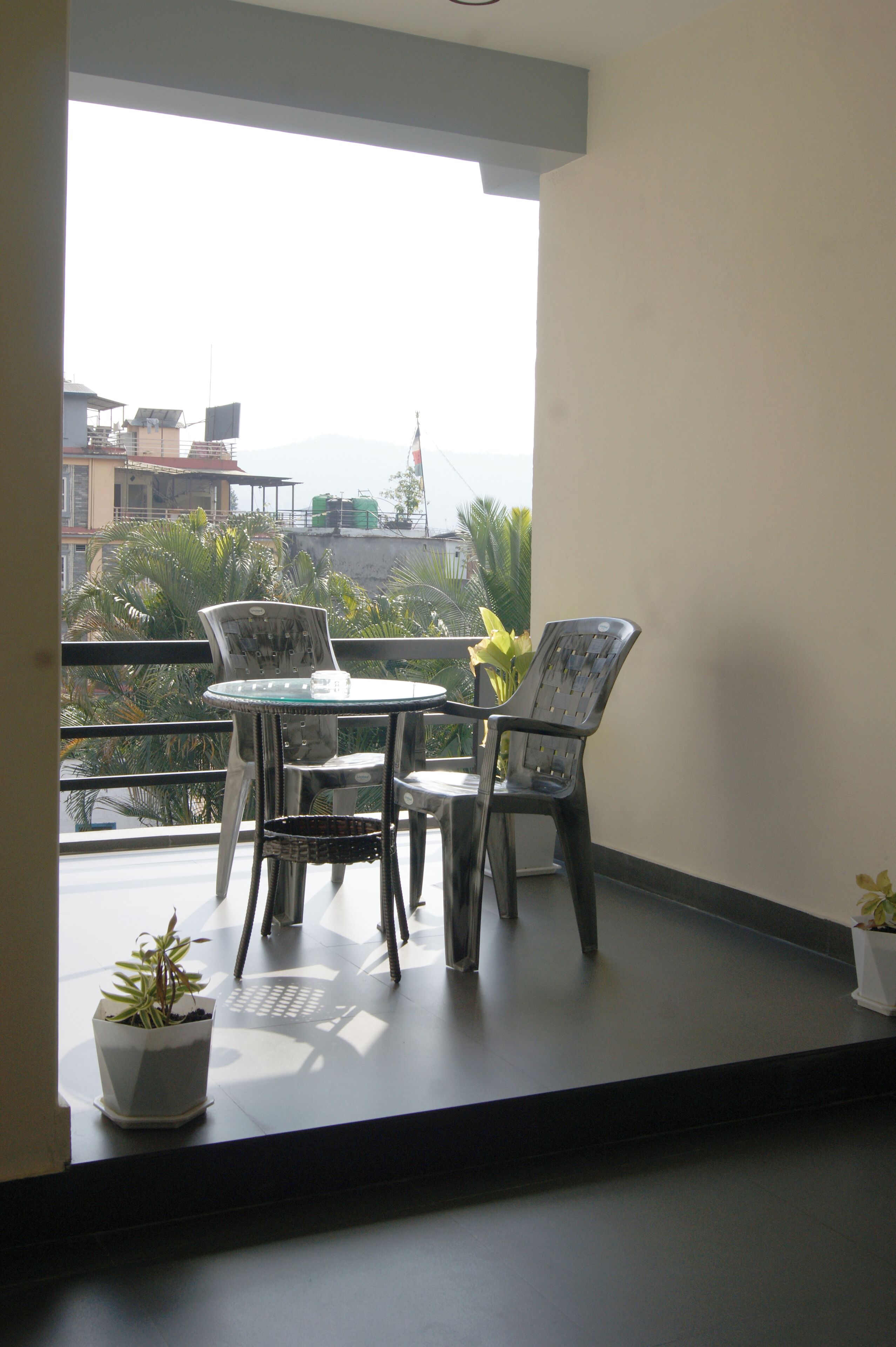 A balcony with a table and chairs, potted plants, and a view of buildings and greenery.