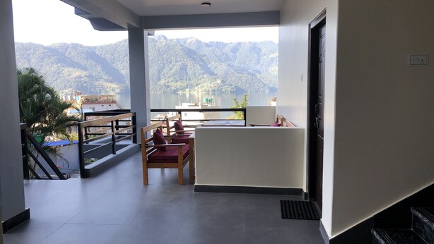 Un balcón con vistas a montañas y un lago, una silla de madera con cojín y una mesita.