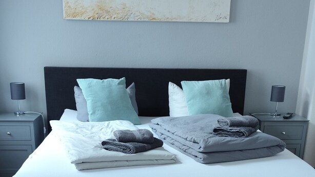 A neatly made bed with pillows and folded towels, a bedside table with a lamp, and a wooden floor.
