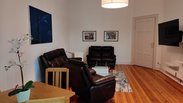 A living room with a brown leather sofa, a wooden coffee table, and a small potted plant. There are two framed pictures on the wall, a door, and a television on a stand. - Vacation apartment Cottbuser City (Cottbus)