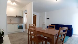 A modern kitchen with white cabinets and a built-in oven, a dining area with a wooden table and chairs, and a blue sofa in the living room.