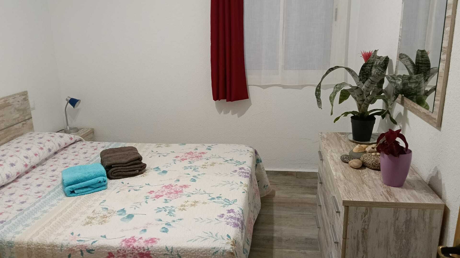 A bedroom with a bed, a nightstand, a dresser, a potted plant, and a window with a red curtain.