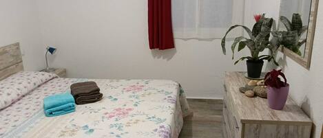 A bedroom with a bed, a nightstand, a dresser, a potted plant, and a window with a red curtain.