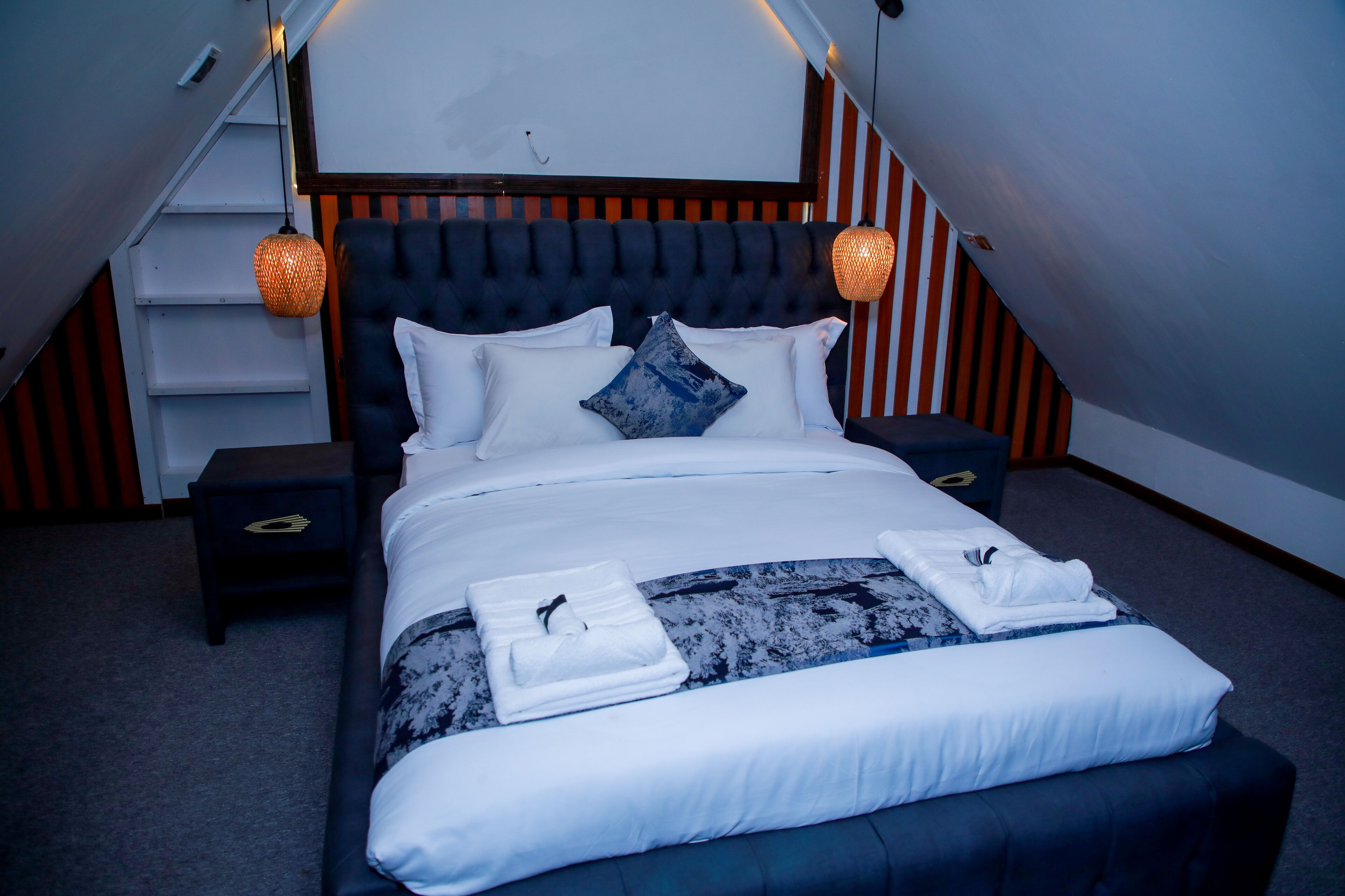 A neatly made bed with white linens and a blue and white patterned throw, two bedside tables with lamps, and a staircase with striped walls.