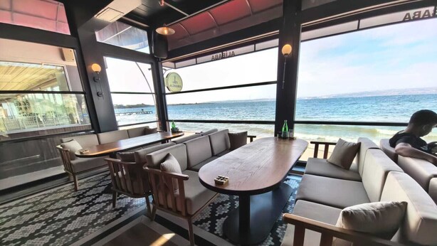 Un interior moderno con un ventanal grande que ofrece vistas al mar, una zona de estar con sofá y sillones, y una mesa de madera.