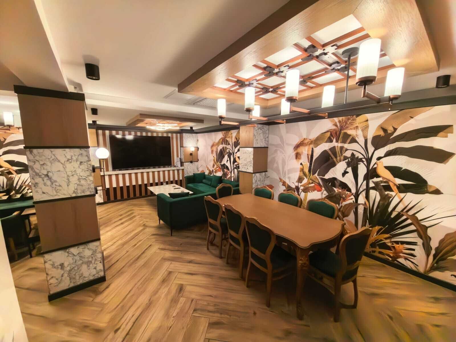 A modern living room with a wooden herringbone floor, a green sofa, a wooden dining table with chairs, a flat-screen TV, and a large wall mural.
