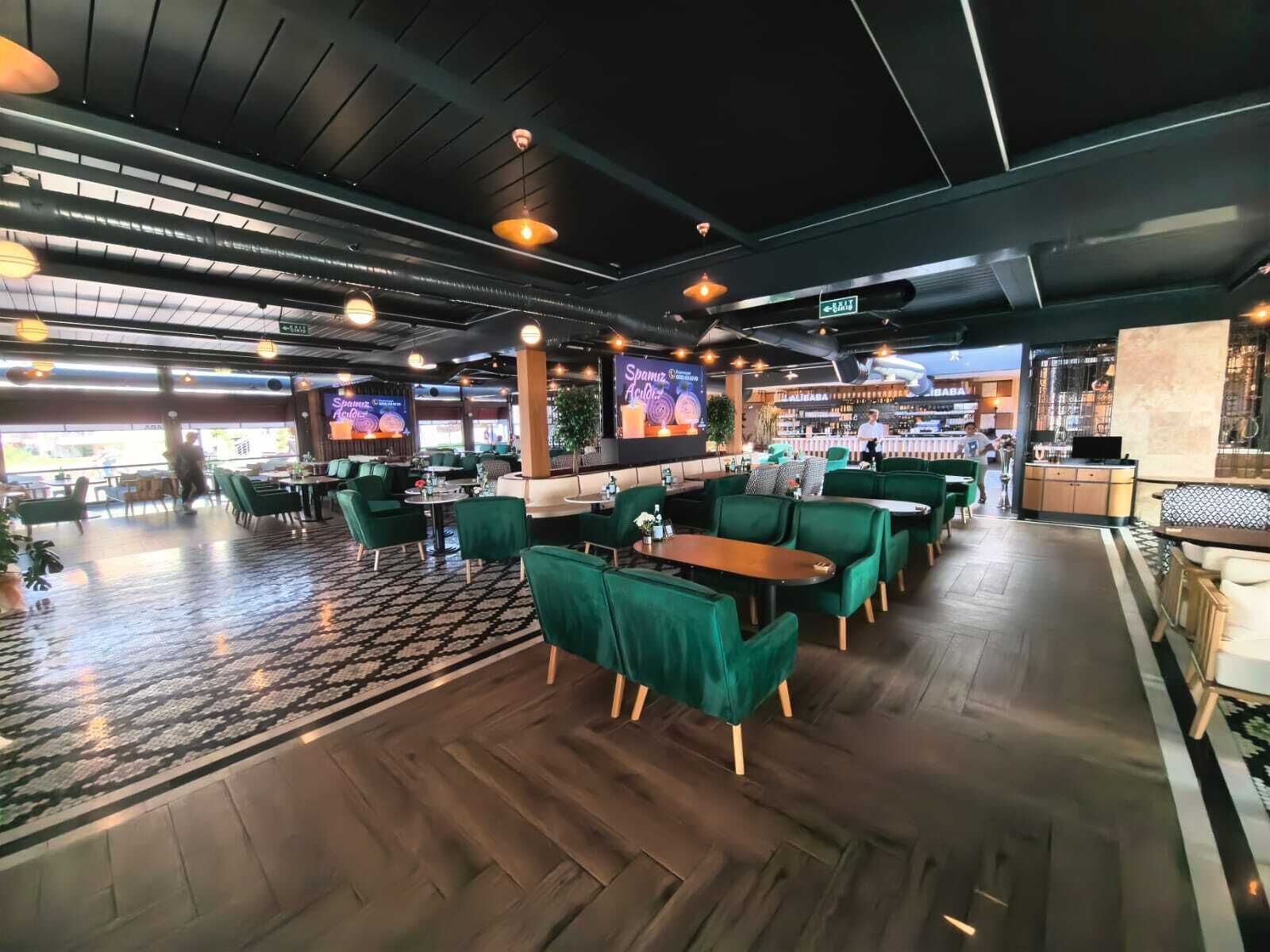 A modern restaurant with green upholstered seating, a patterned floor, and a bar area in the background.