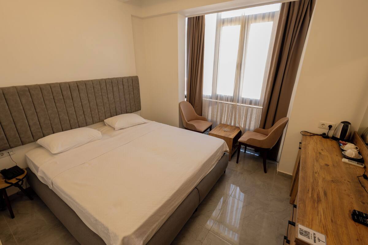 A hotel room with a large bed, a desk with a chair, a small table, and a window with curtains.
